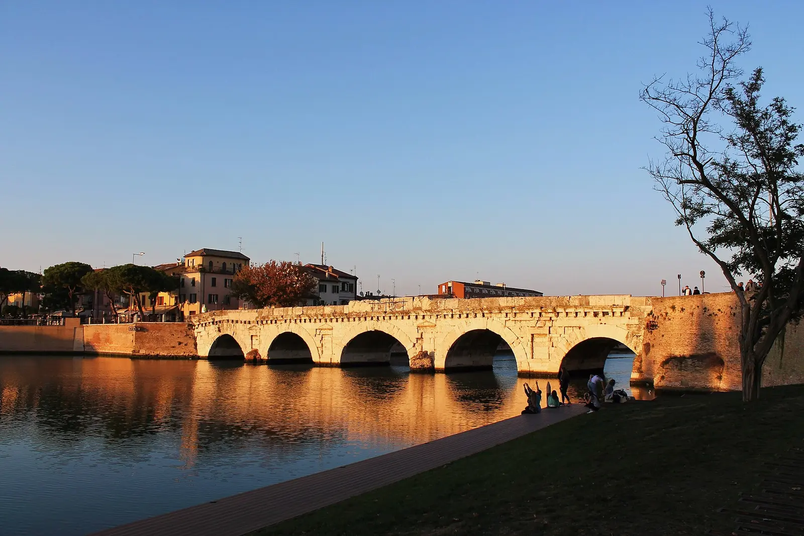Il Ponte di Tiberio di Rimini nella luce dorata del tramonto, con le cinque arcate sul canale e un prato in primo piano