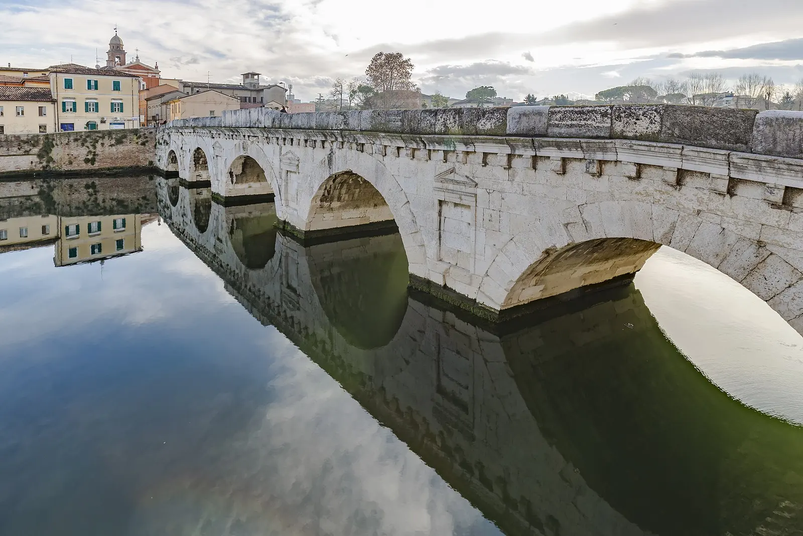 Les arches du Ponte di Tiberio à Rimini se reflètent dans l'eau calme, avec les maisons colorées en arrière-plan