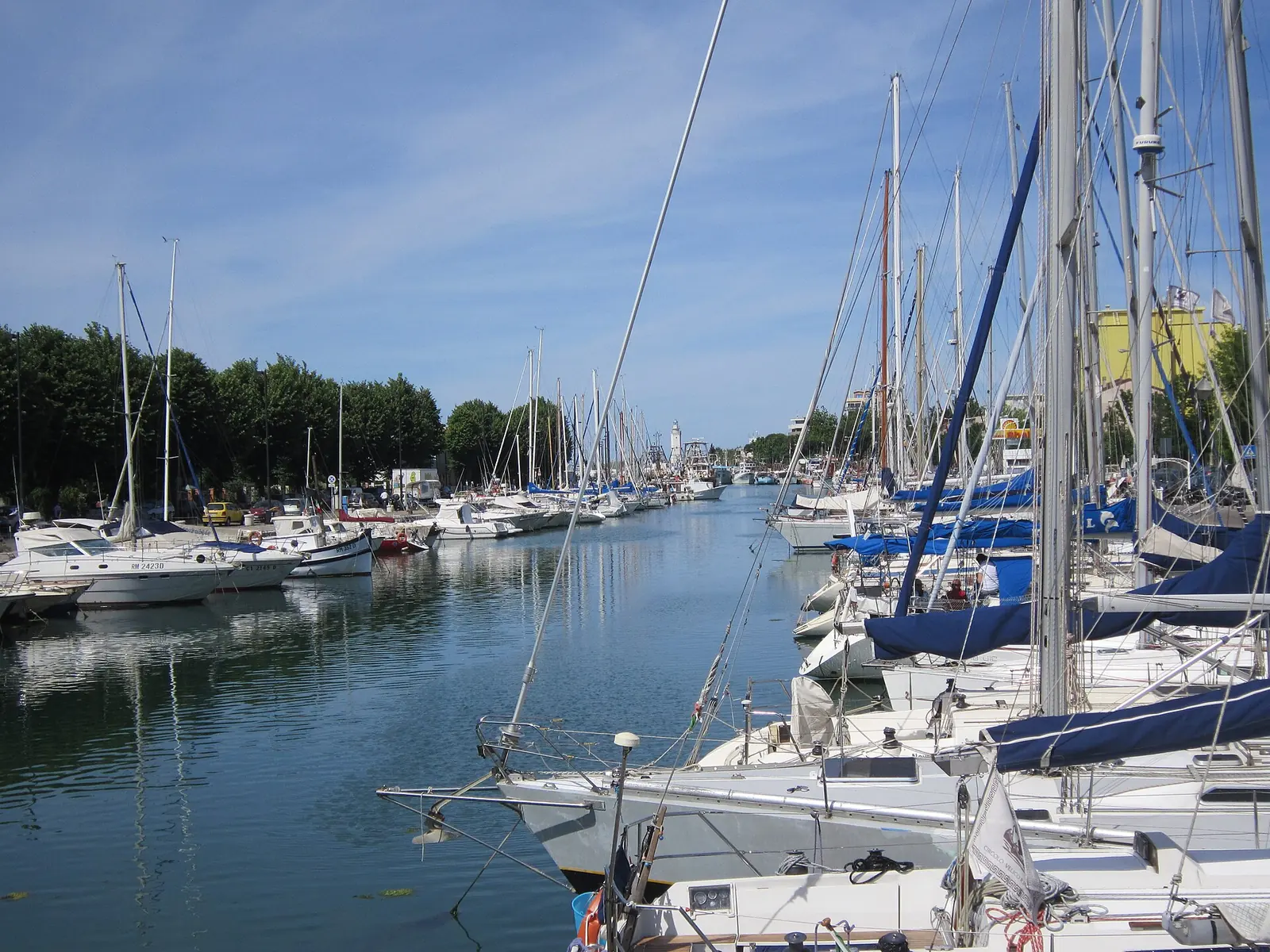 Il porto canale di Rimini con le barche a vela ormeggiate lungo la banchina e il faro in fondo al canale