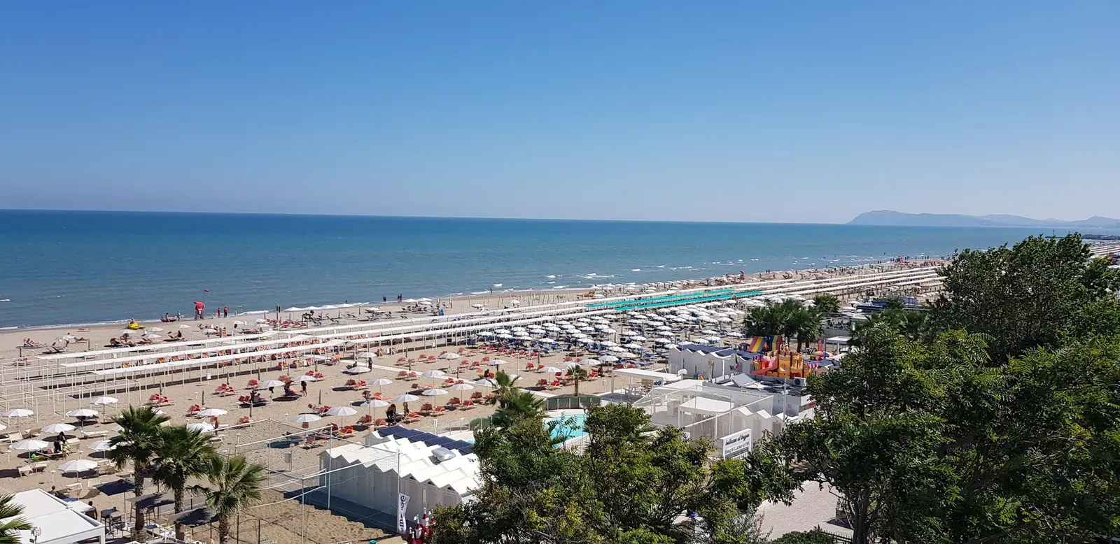 Die Küste von Riccione von oben mit Sonnenschirmreihen, weißen Hütten und blauem Meer bis zur Landzunge