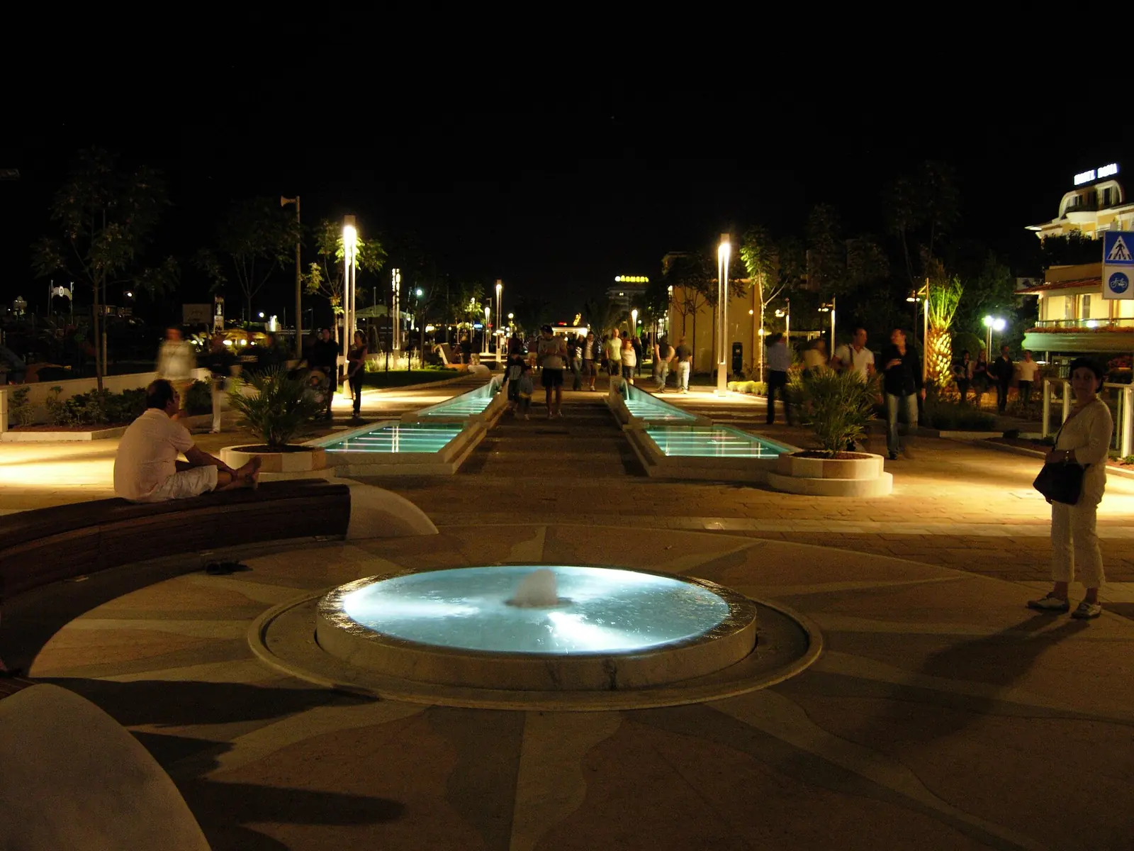 Die Strandpromenade von Riccione am Abend mit beleuchteten Wasserbecken, Laternen und Passanten in der Ferne