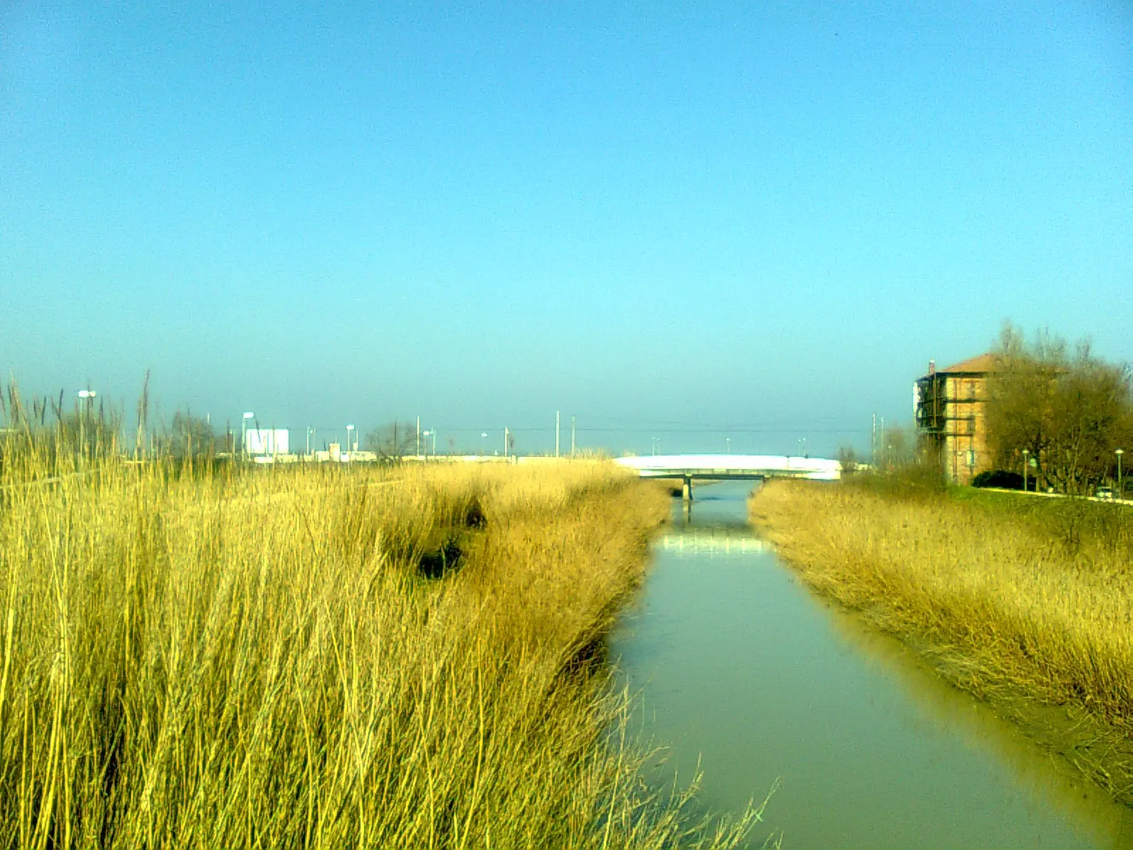 Der Marano in Riccione zwischen zwei Ufern mit hohem Gras, mit einer weißen Brücke und dem Meer in der Ferne