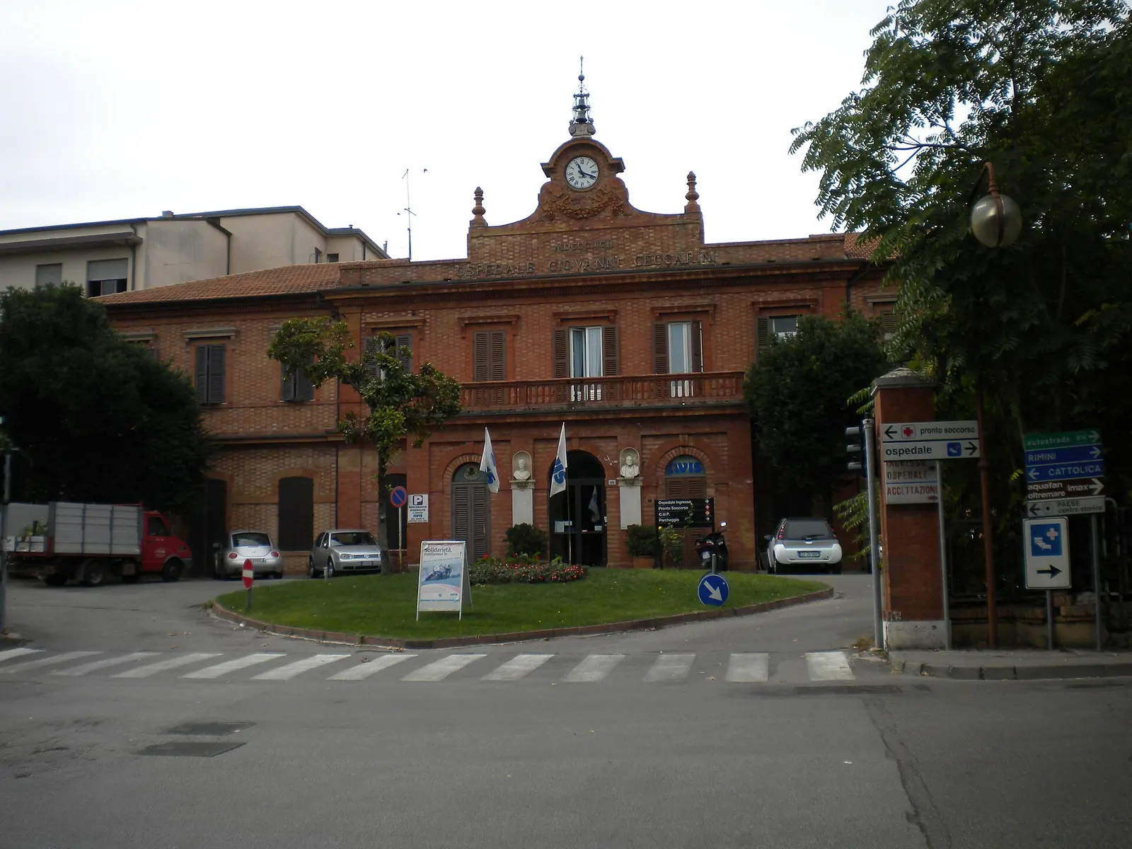 La façade en brique de l'Ospedale Ceccarini de Riccione, avec l'horloge au sommet et le parterre rond devant l'entrée