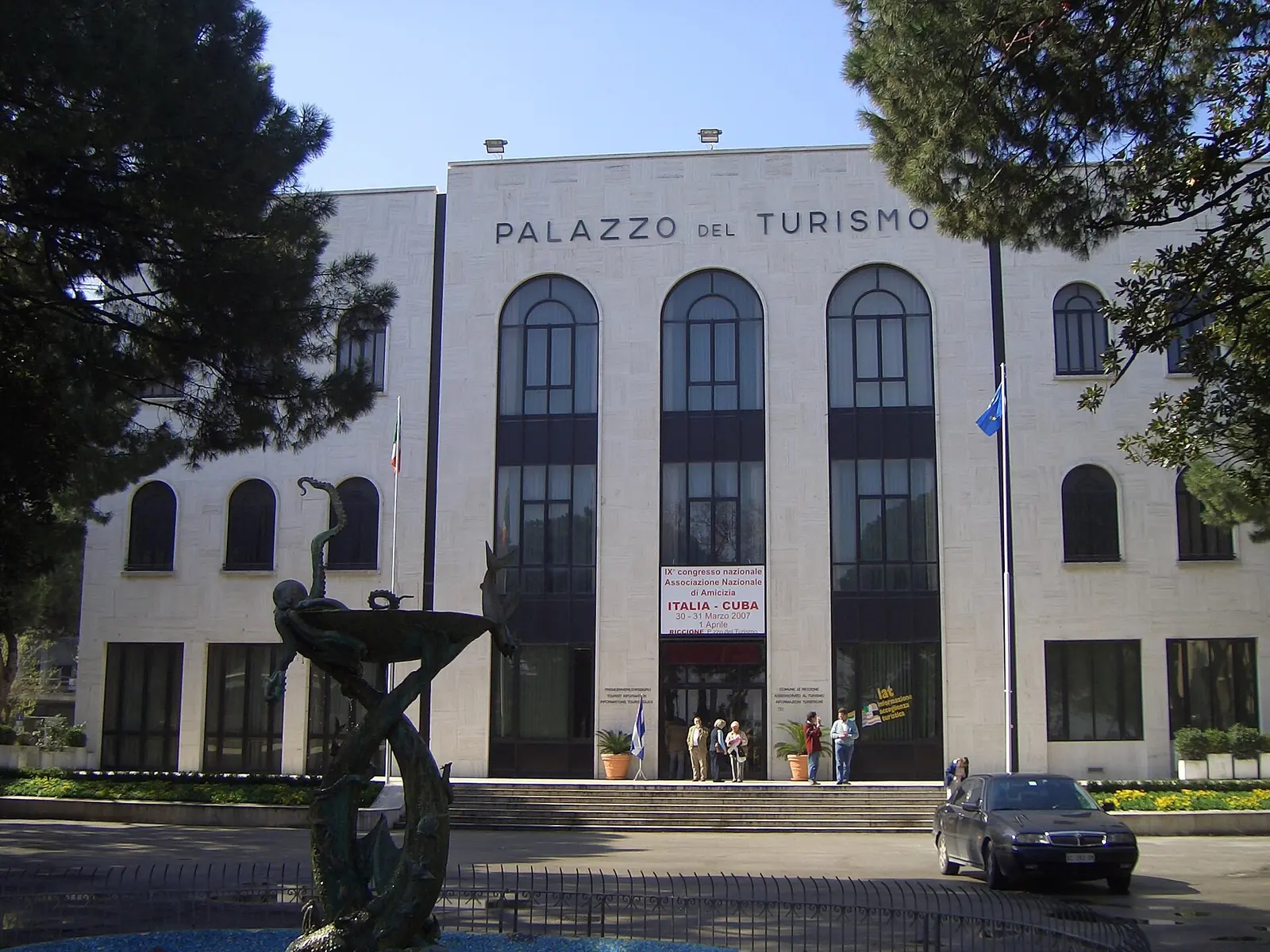 Le Palazzo del Turismo de Riccione, bâtiment clair aux grandes fenêtres en arc, parmi les pins et avec une fontaine au premier plan