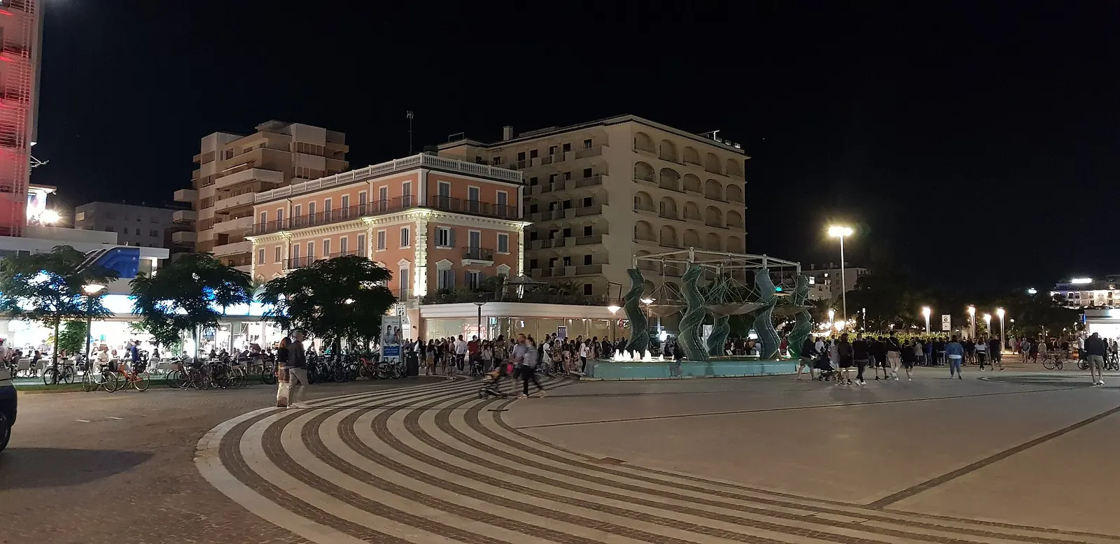 Piazzale Roma a Riccione di sera, in fondo a viale Ceccarini, con la fontana illuminata e la folla del passeggio