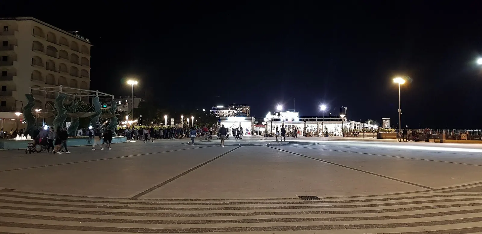 Piazzale Roma in Riccione in the evening, with the illuminated fountain, distant passers-by and the seafront in the background