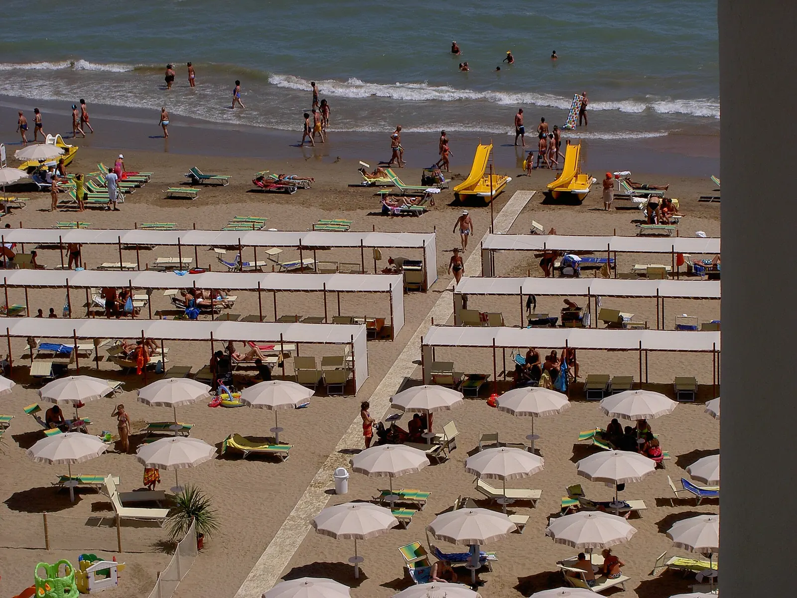 A Riccione beach seen from above in summer, with rows of white beach tents and parasols and bathers by the sea