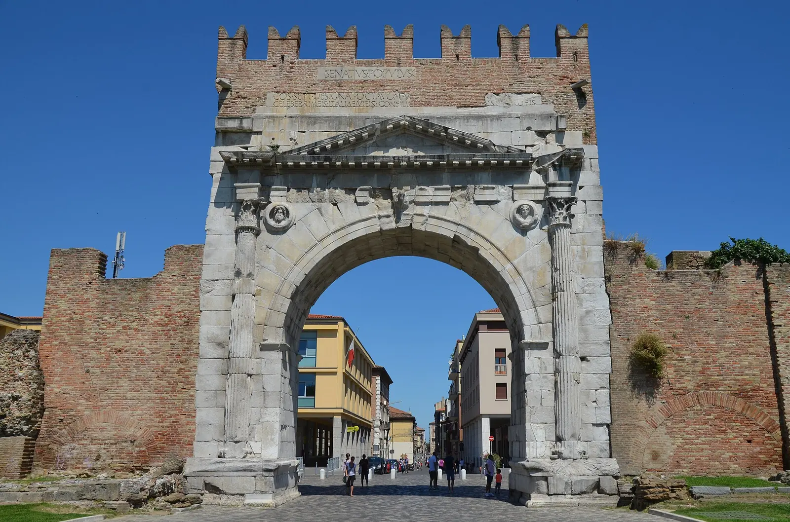 L'Arco di Augusto di Rimini visto di fronte, con la merlatura in laterizio, le mura ai lati e il corso sullo sfondo