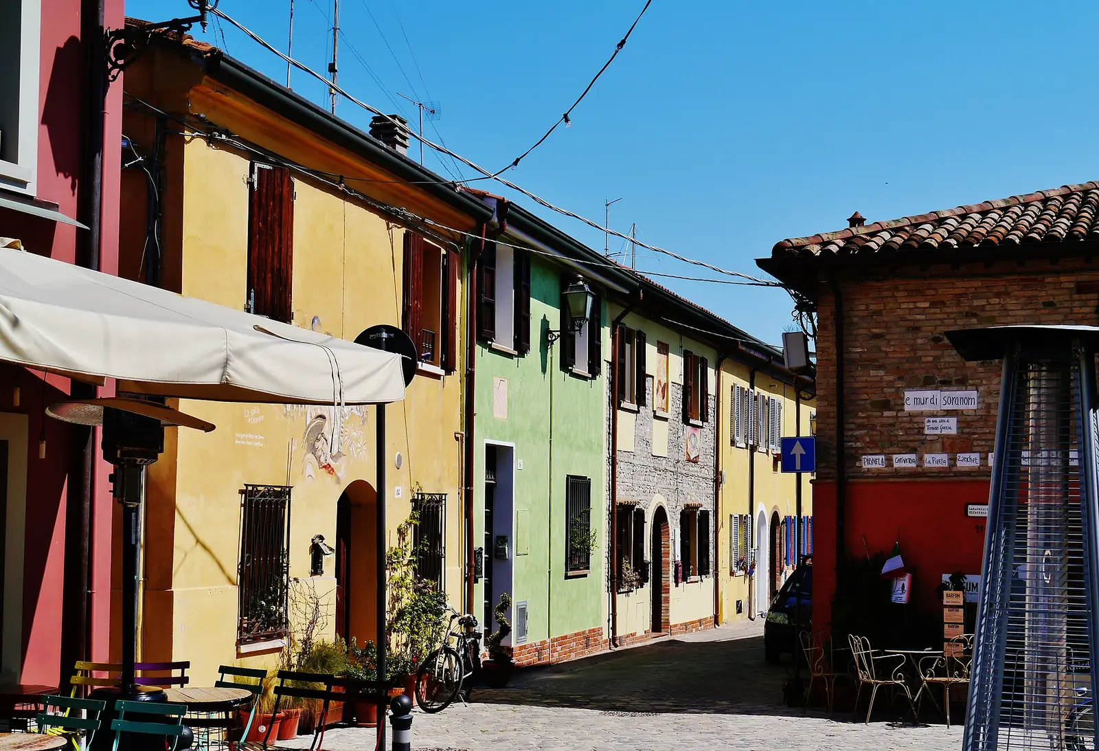 Una via del Borgo San Giuliano a Rimini, con case basse gialle, verdi e rosse, tavolini all'aperto e selciato
