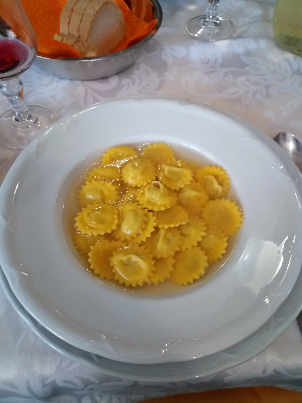 Ein Teller Cappelletti in Brühe auf einer hellen Tischdecke mit einem Glas Rotwein und einem Brotkorb