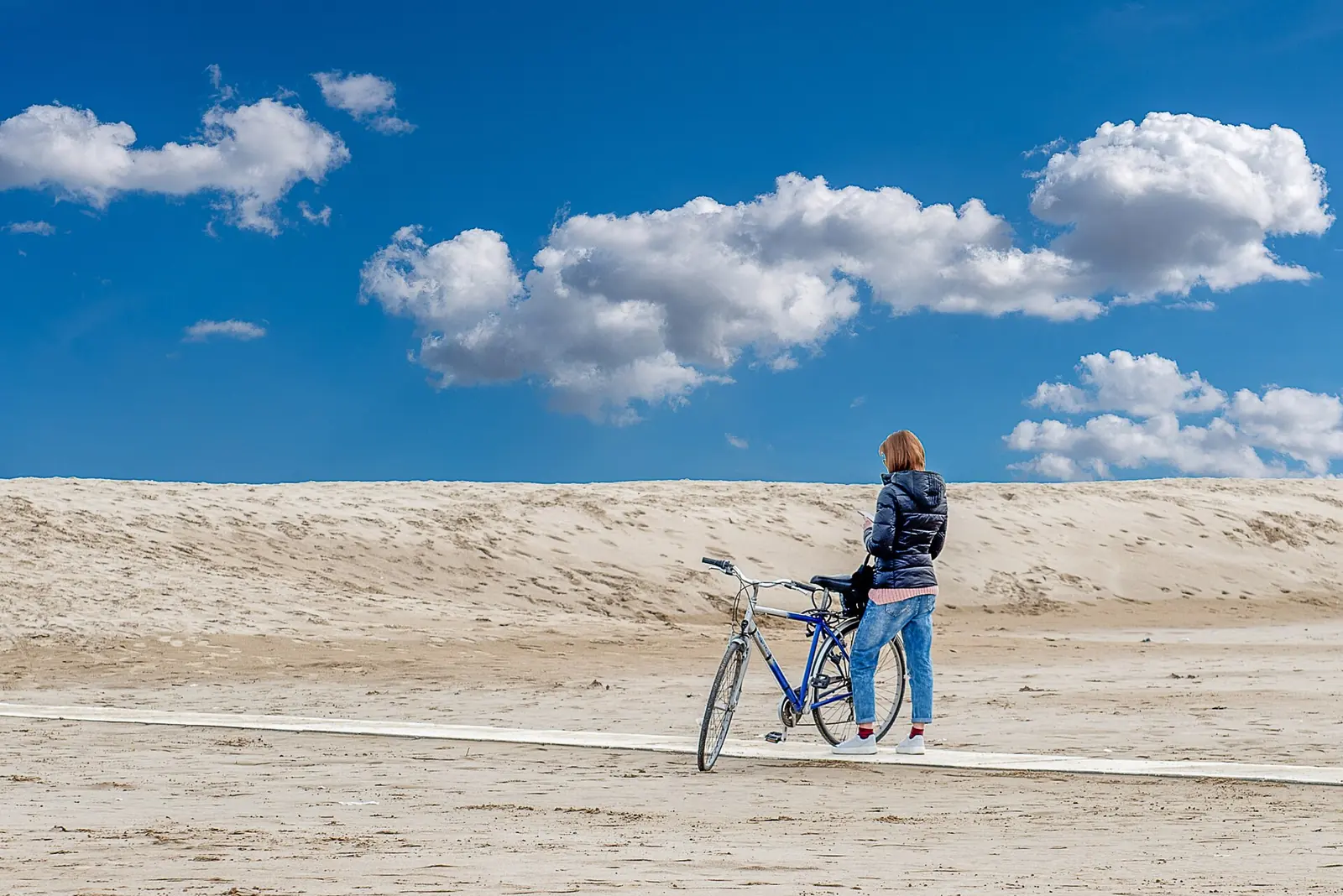 A woman seen from behind with a blue bicycle on a narrow pale walkway, in front of a dune and cloudy sky