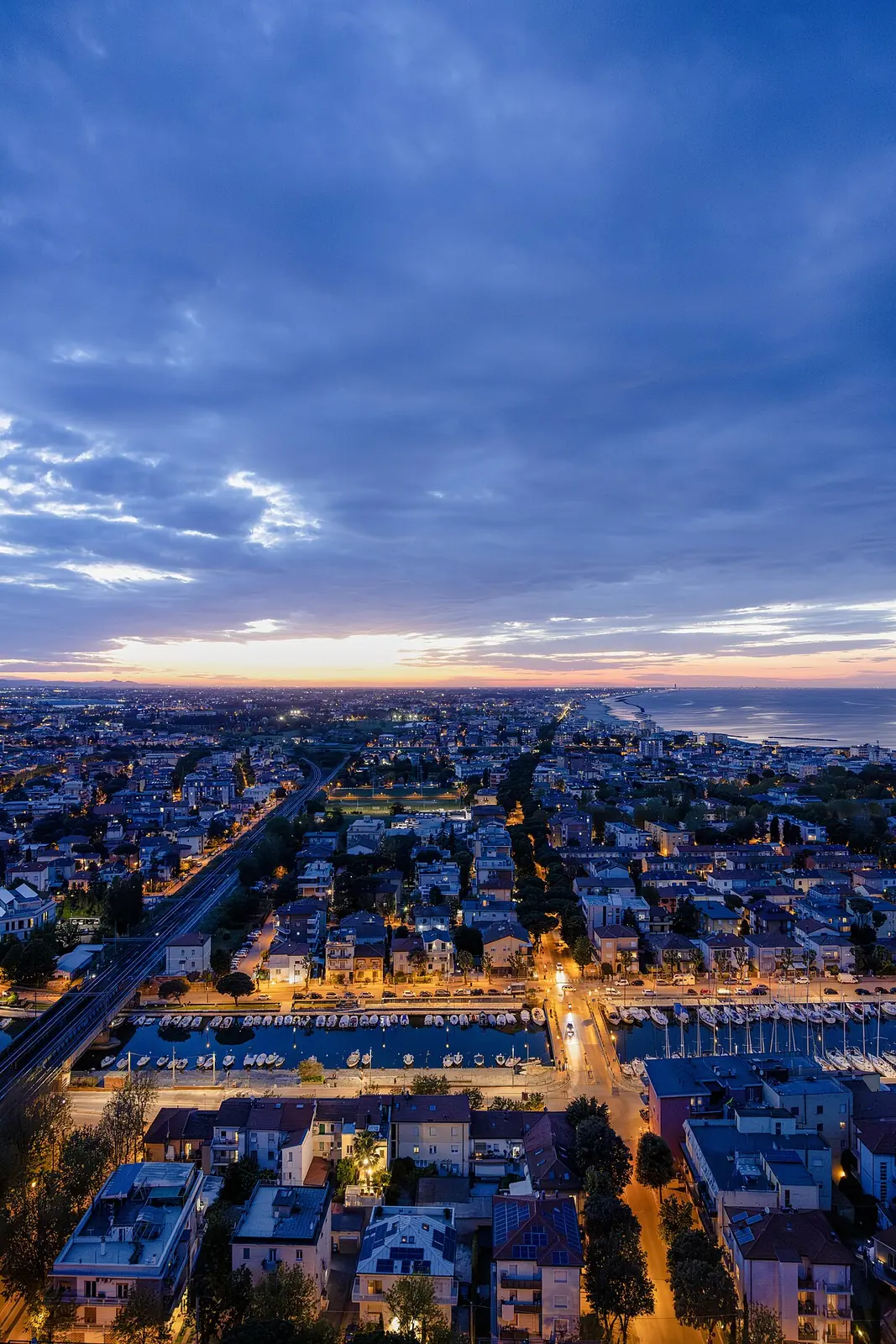 Rimini at dusk seen from above, with the canal harbour full of boats, the railway and coast on the horizon