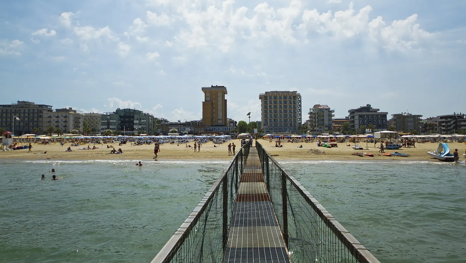 A jetty over the sea in Rimini, with the beach, parasols and seafront buildings in the background