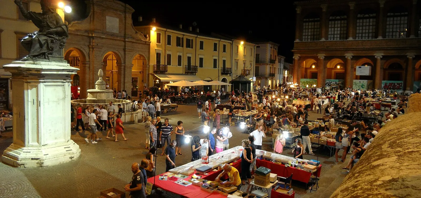 Der Abendmarkt auf der Piazza Cavour in Rimini mit beleuchteten Ständen, Passanten und der Statue auf dem Sockel