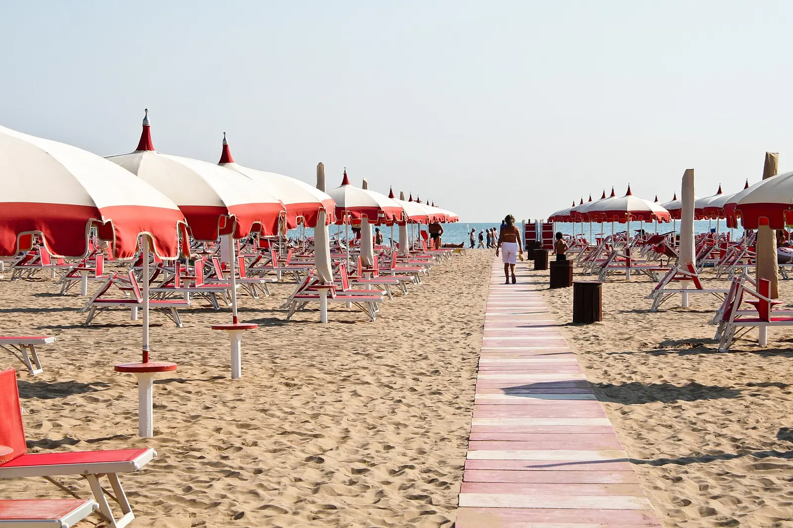 Der Strand von Rimini im Sommer mit weißen und roten Sonnenschirmen, Liegenreihen und einem Holzsteg zum Meer