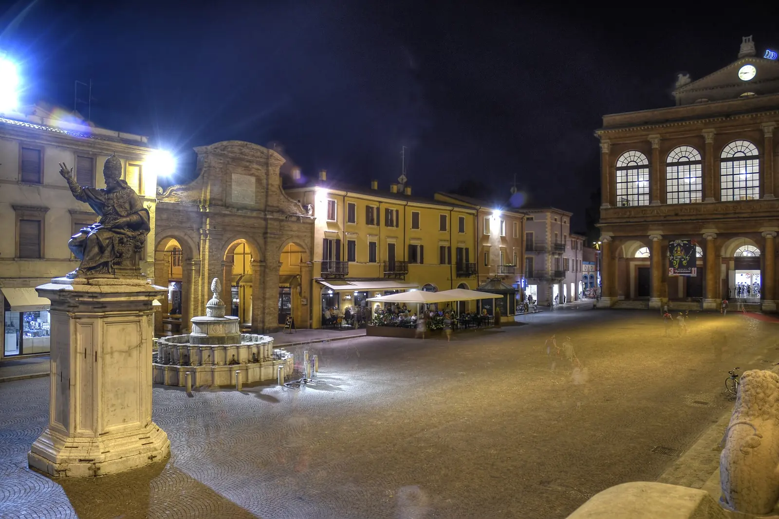 Piazza Cavour a Rimini di notte, con la statua di Paolo V, la fontana, i palazzi illuminati e il Teatro Galli sulla destra