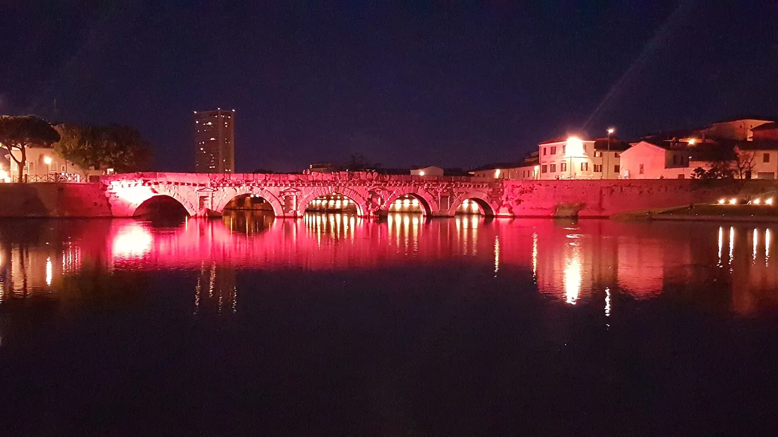 Il Ponte di Tiberio di Rimini illuminato di rosa di sera, con il riflesso rosso nell'acqua del Marecchia