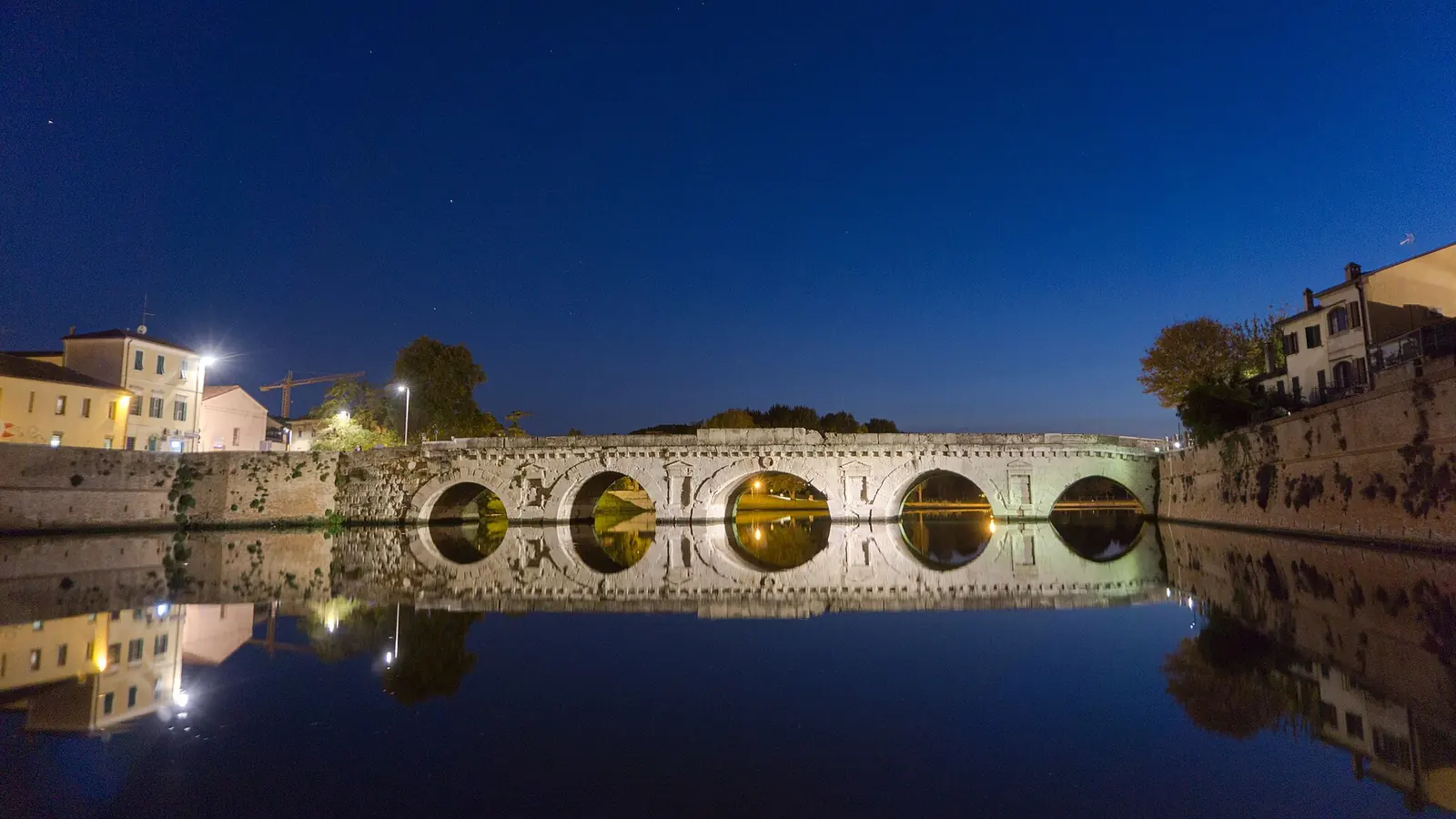 Il Ponte di Tiberio di Rimini illuminato di notte, con le arcate specchiate nell'acqua del porto canale