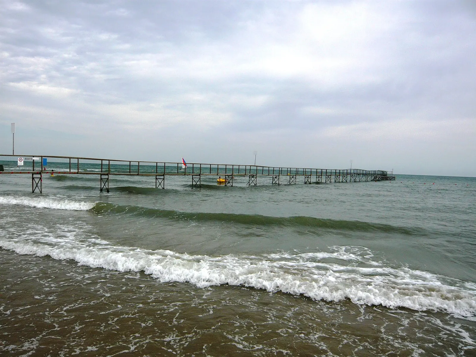 A long iron jetty extending over the sea in Rimini, with waves at the waterline and an overcast sky