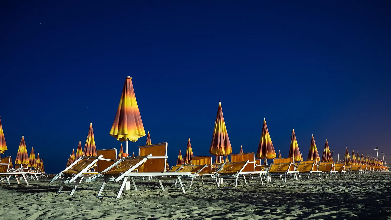 La spiaggia di Rimini di notte, con gli ombrelloni chiusi illuminati di giallo e i lettini in fila sotto un cielo blu scuro
