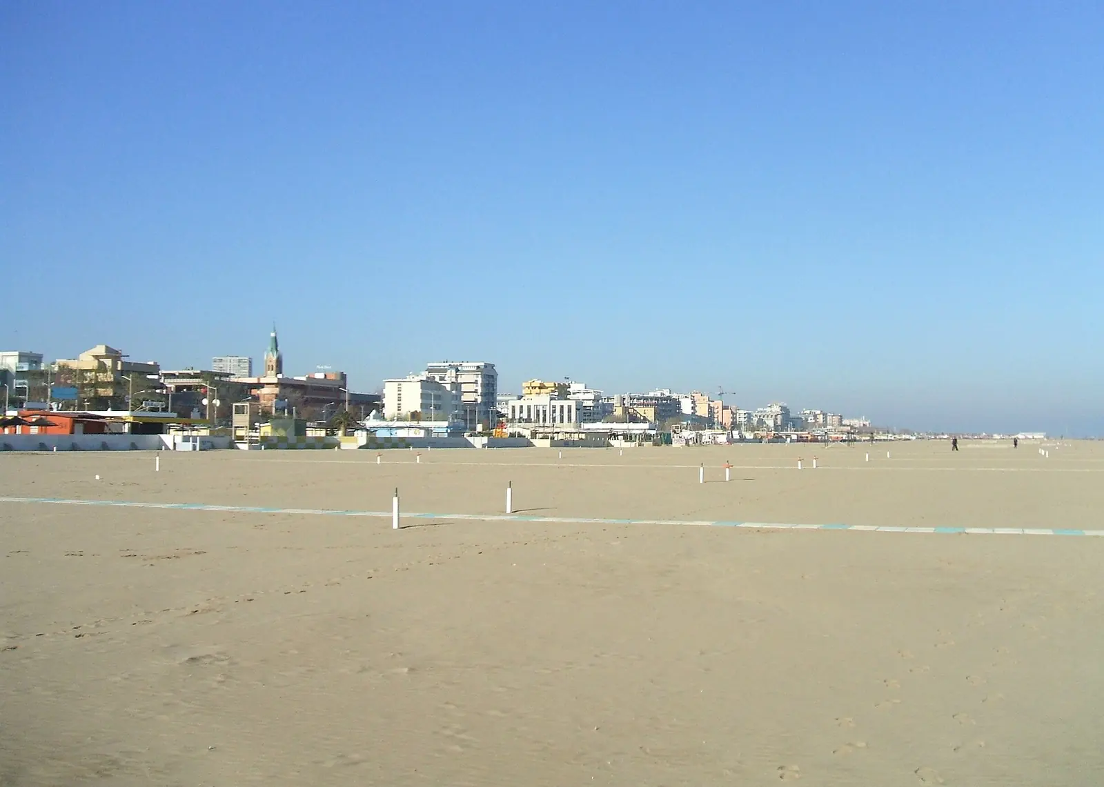 L'arenile di Rimini fuori stagione, vuoto e ampio, con una passerella chiara e i palazzi del lungomare in lontananza