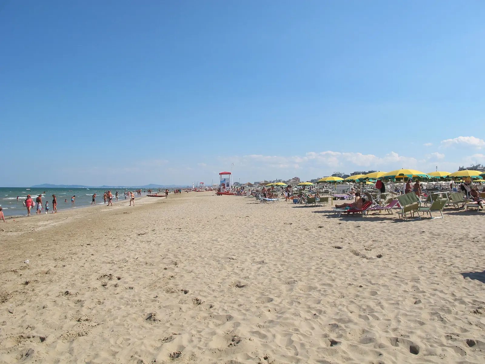 L'ampia spiaggia sabbiosa di Rimini in estate, con il bagnasciuga a sinistra e file di ombrelloni gialli a destra