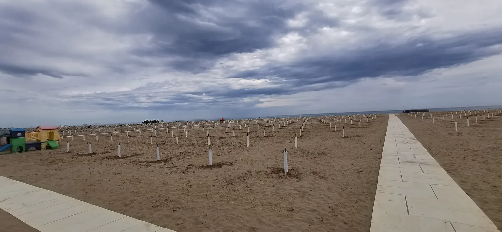 Ein Strand von Rimini außerhalb der Saison mit Sonnenschirmstangen, einem hellen Steg und einem Spielbereich links