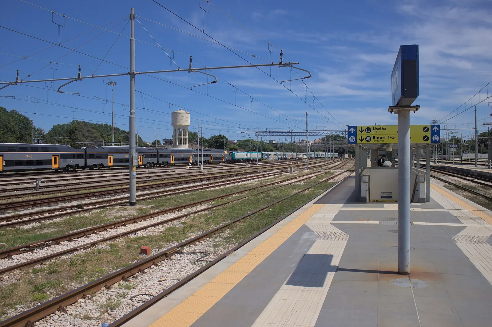 A platform at Rimini station with the yellow tactile guidance strip, signs for exits and platforms, and trains in the background