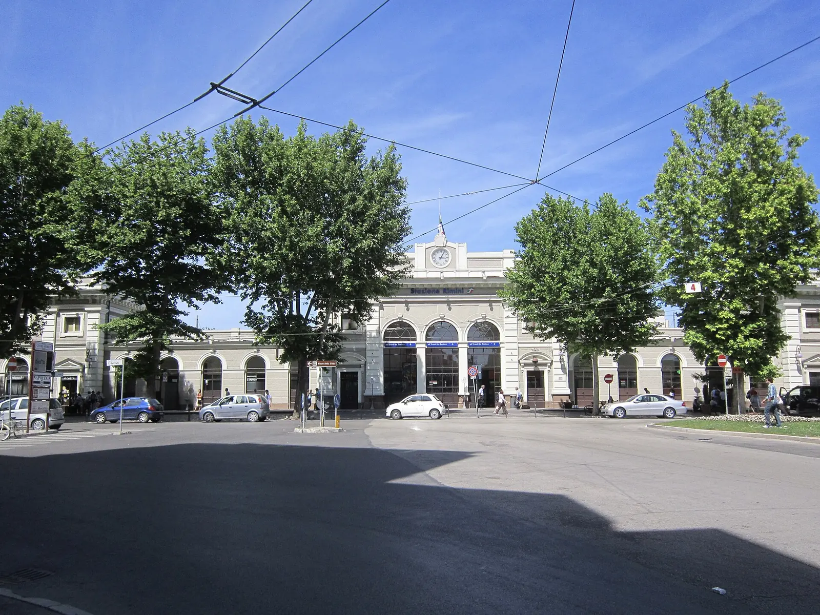 La facciata della stazione ferroviaria di Rimini con l'orologio sul frontone, gli alberi del piazzale e le auto parcheggiate