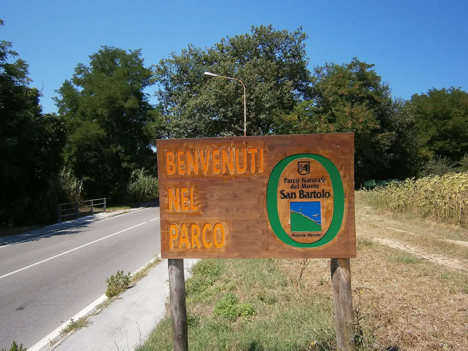Il cartello di legno Benvenuti nel Parco con lo stemma del Parco del Monte San Bartolo, accanto a una strada fra gli alberi