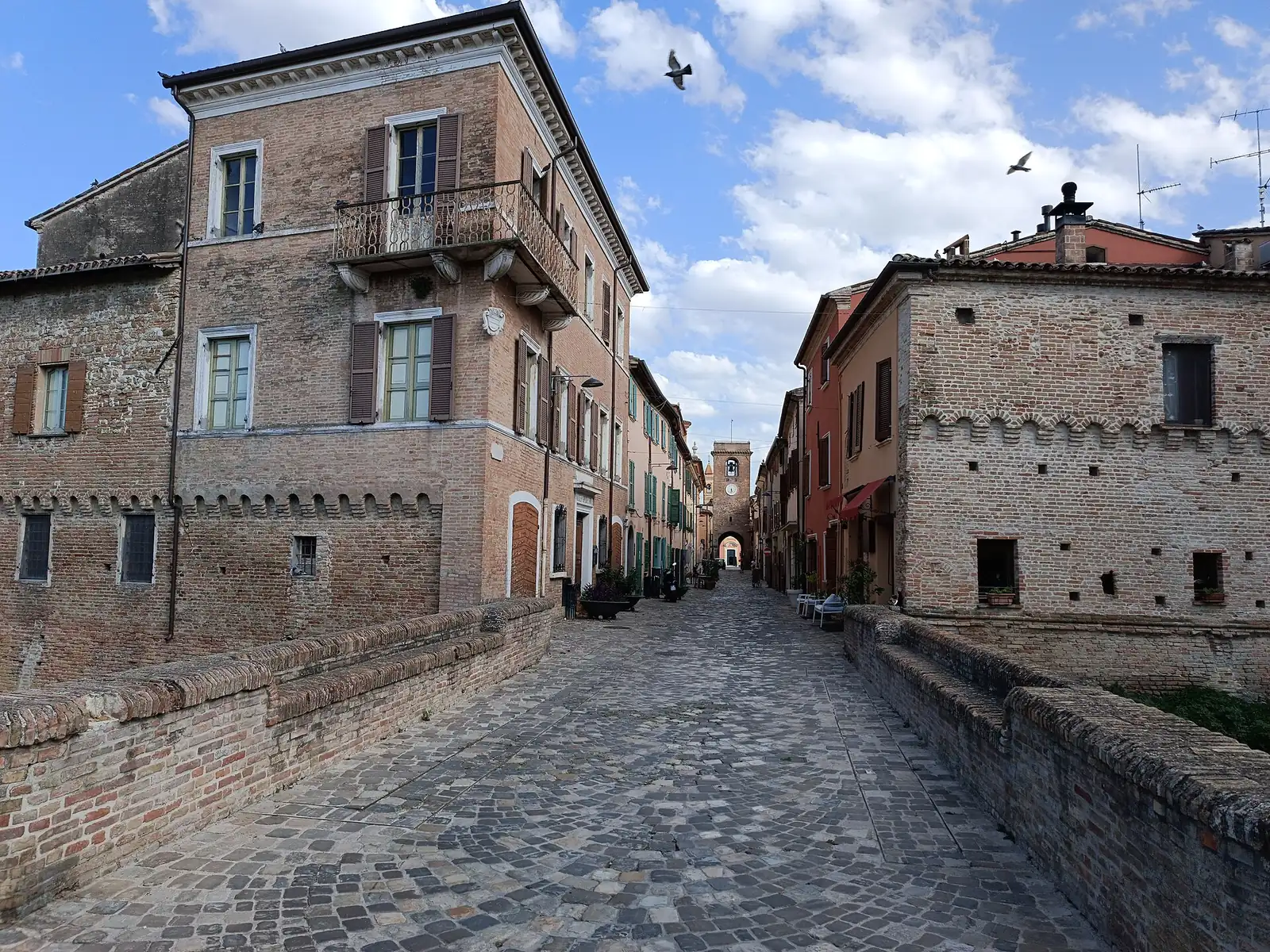 The cobbled street of San Giovanni in Marignano among brick houses, with the clock tower at the far end