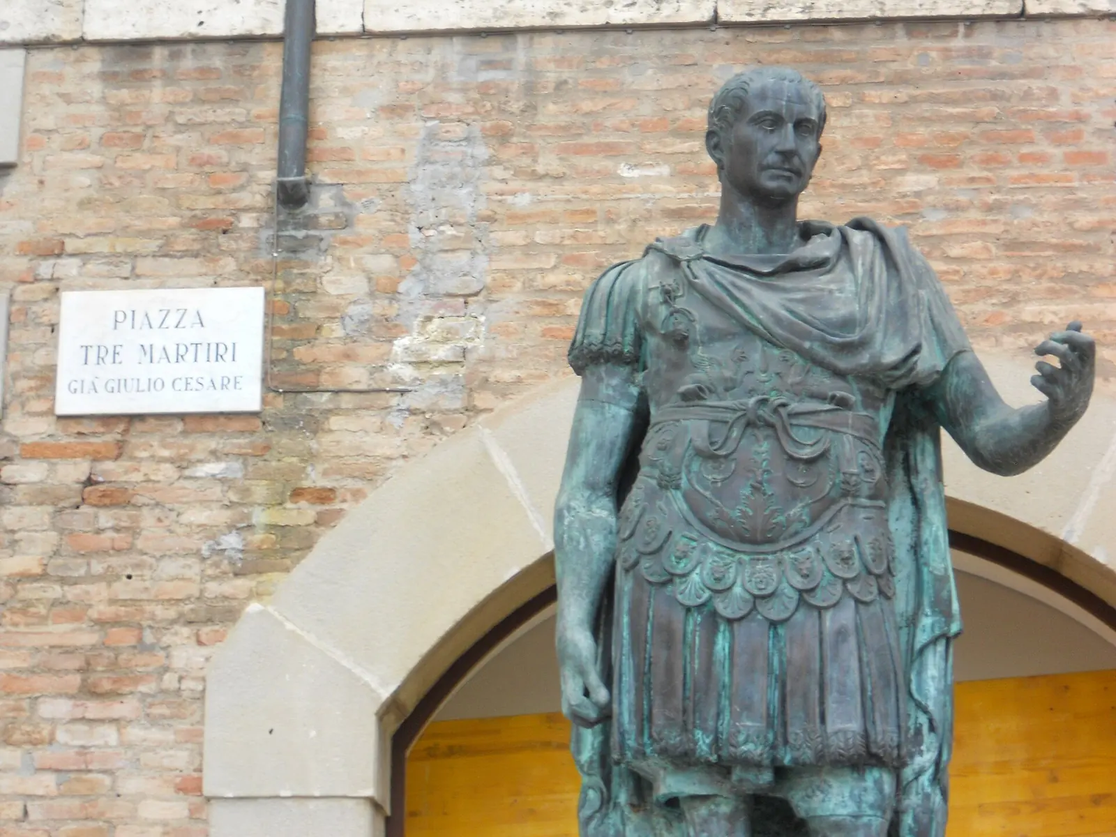 Die Bronzestatue von Giulio Cesare auf der Piazza Tre Martiri in Rimini neben der Tafel „Piazza Tre Martiri già Giulio Cesare“