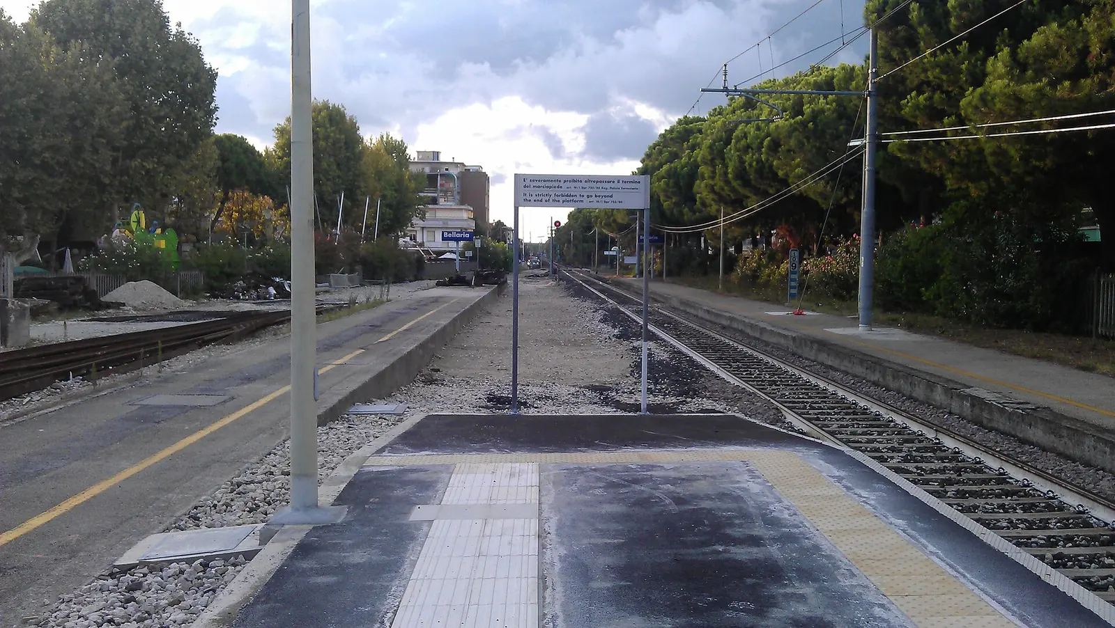 La banchina della stazione di Bellaria verso Ravenna, con un cartello di sicurezza sui binari e i pini sulla destra