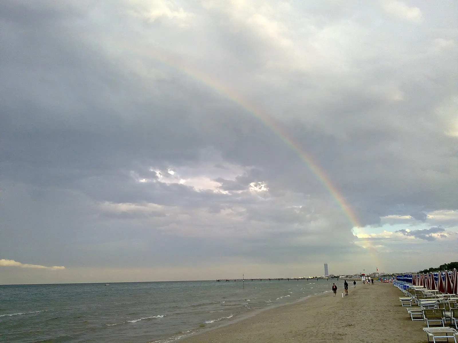 Un arcobaleno sul mare davanti alla spiaggia di Tagliata di Cervia, con alcune persone a passeggio sulla riva