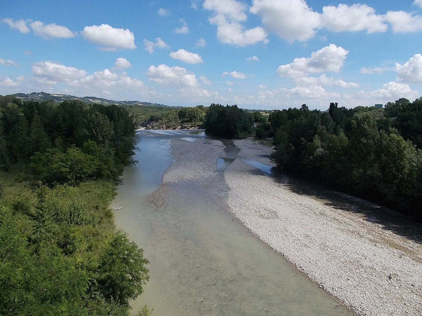 Il letto ghiaioso del fiume Marecchia visto dall'alto di un ponte, con gli alberi sulle rive e le colline sullo sfondo