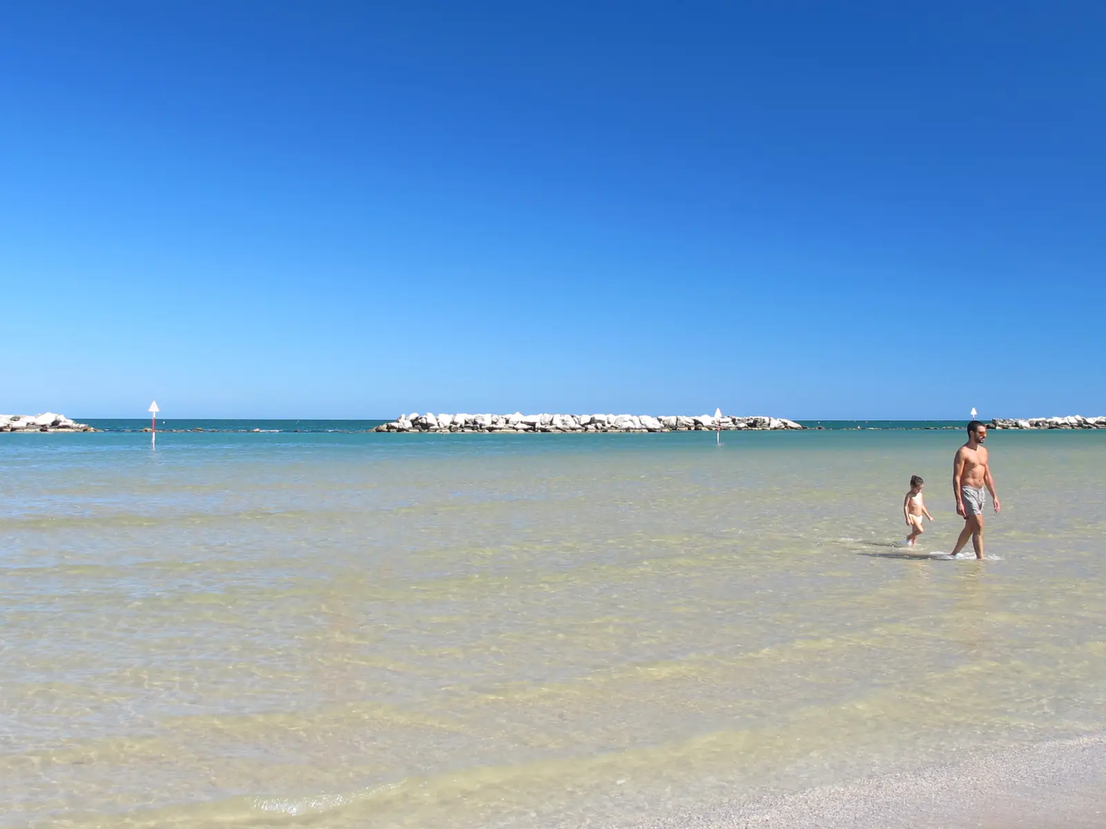Due persone lontane nell'acqua bassa e trasparente di Bellaria Igea Marina, con le scogliere all'orizzonte