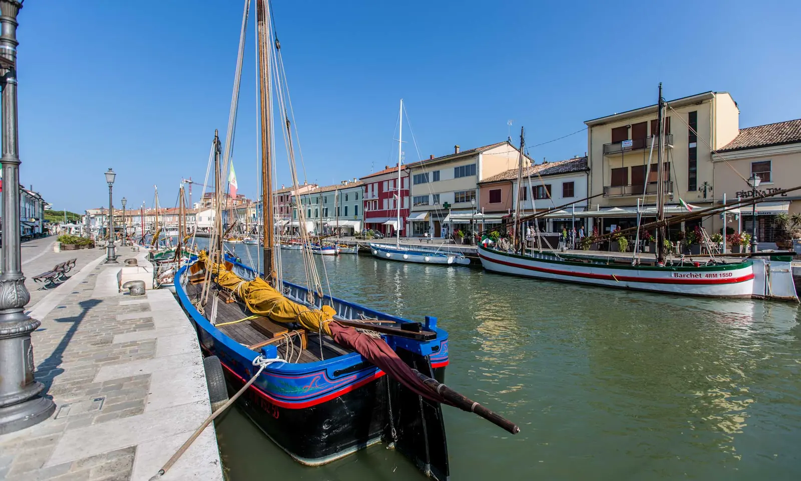 Il porto canale di Cesenatico con un bragozzo blu in primo piano, la banchina lastricata e le case colorate