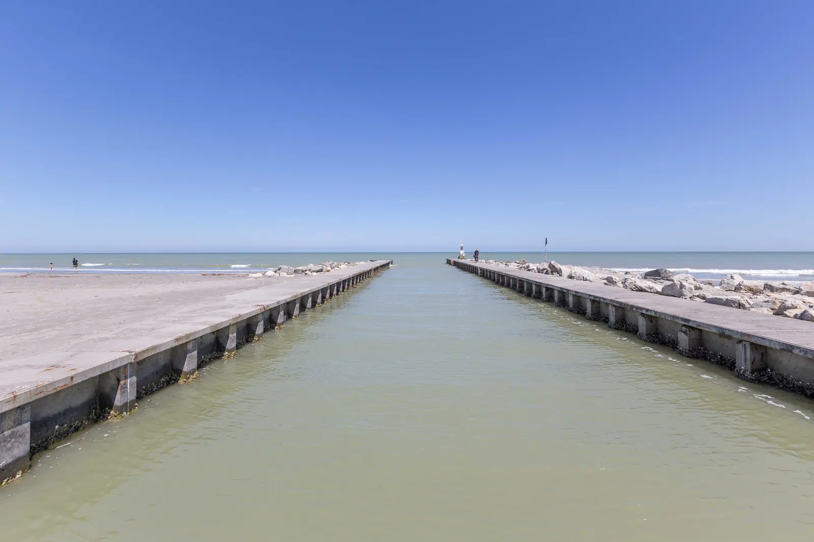 Le due banchine di cemento di un canale che sbocca in mare, con la spiaggia e gli scogli ai lati