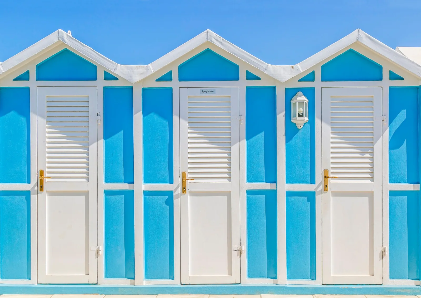 Tre cabine da spiaggia azzurre e bianche a Riccione, con il tetto a punta e le porte a persiana sotto il cielo sereno