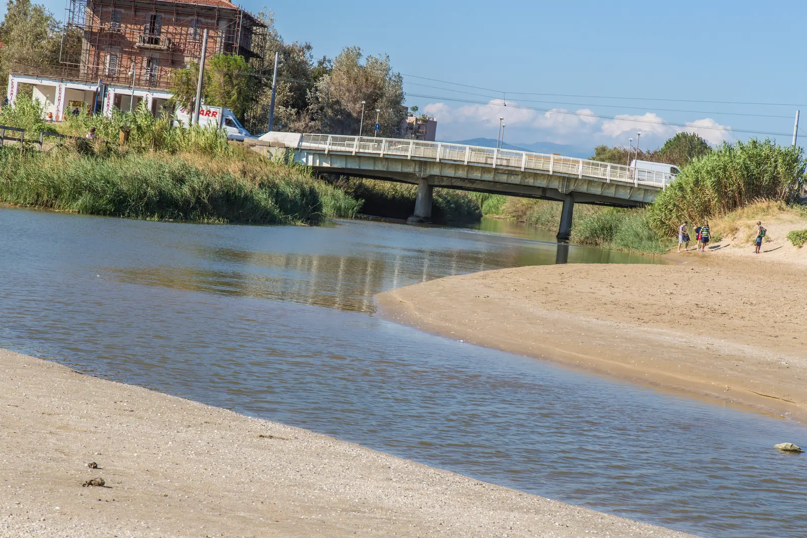 Il torrente Marano a Riccione con le rive di canne, il ponte ciclopedonale bianco e una casa in mattoni sullo sfondo