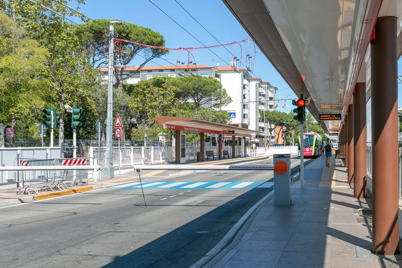 La corsia riservata del Metromare a Riccione con la pensilina rossa della fermata Ceccarini, un filobus in arrivo e i pini