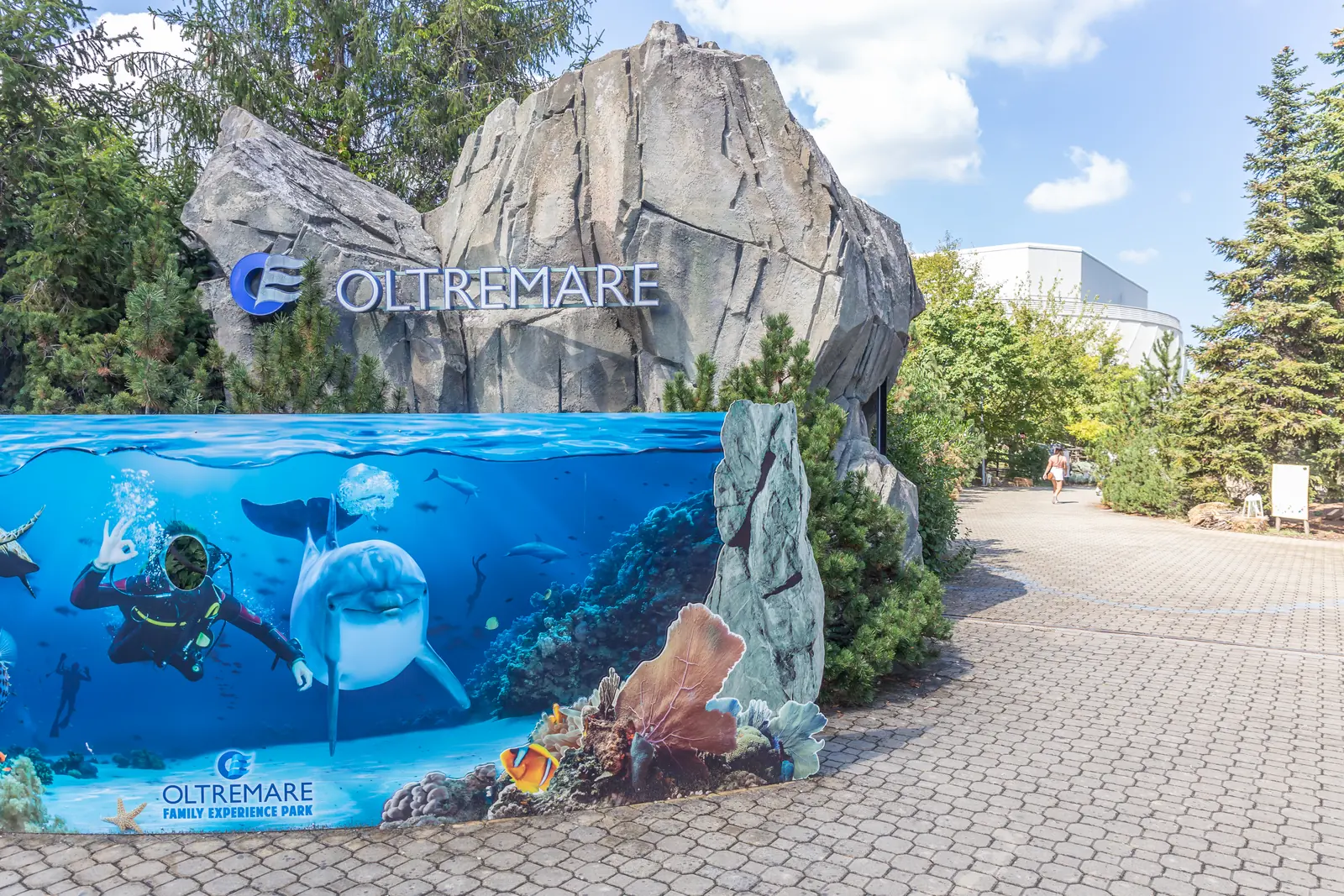 L'ingresso di Oltremare con il nome del parco sulla roccia artificiale, il pannello dipinto con un delfino e un sub e il viale fra gli abeti