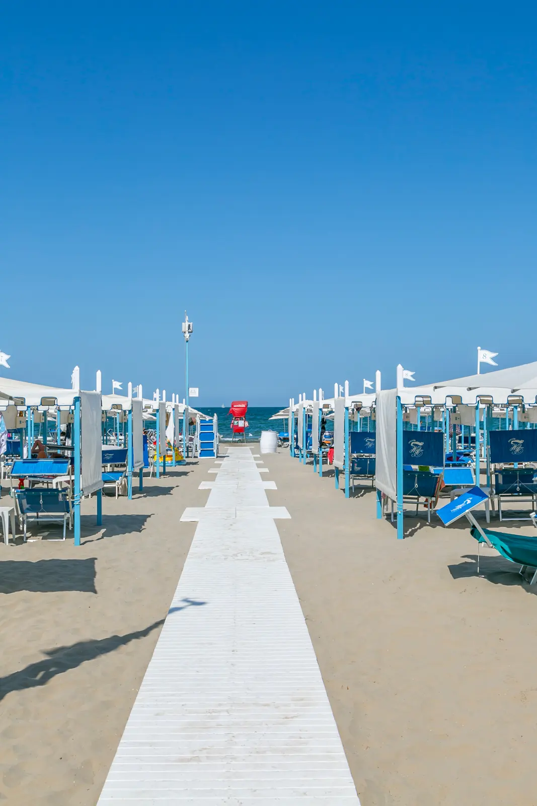 Una passerella bianca sulla sabbia fra due file di ombrelloni, diritta verso il mare