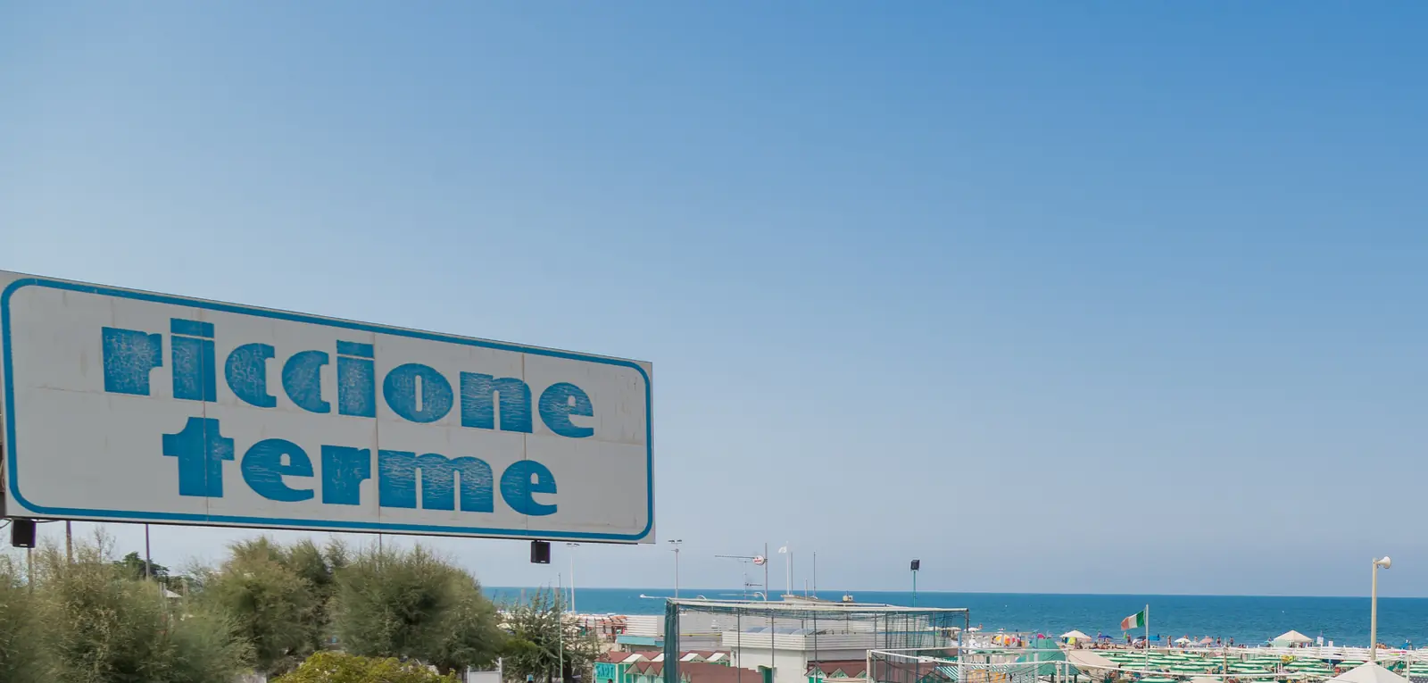 L'insegna «riccione terme» sopra il verde, con il mare e la spiaggia sullo sfondo