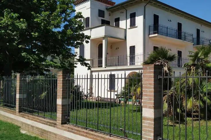 Villa Mussolini a Riccione, edificio bianco a tre piani con la torretta e i balconi in ferro, dietro la recinzione in mattoni e il prato