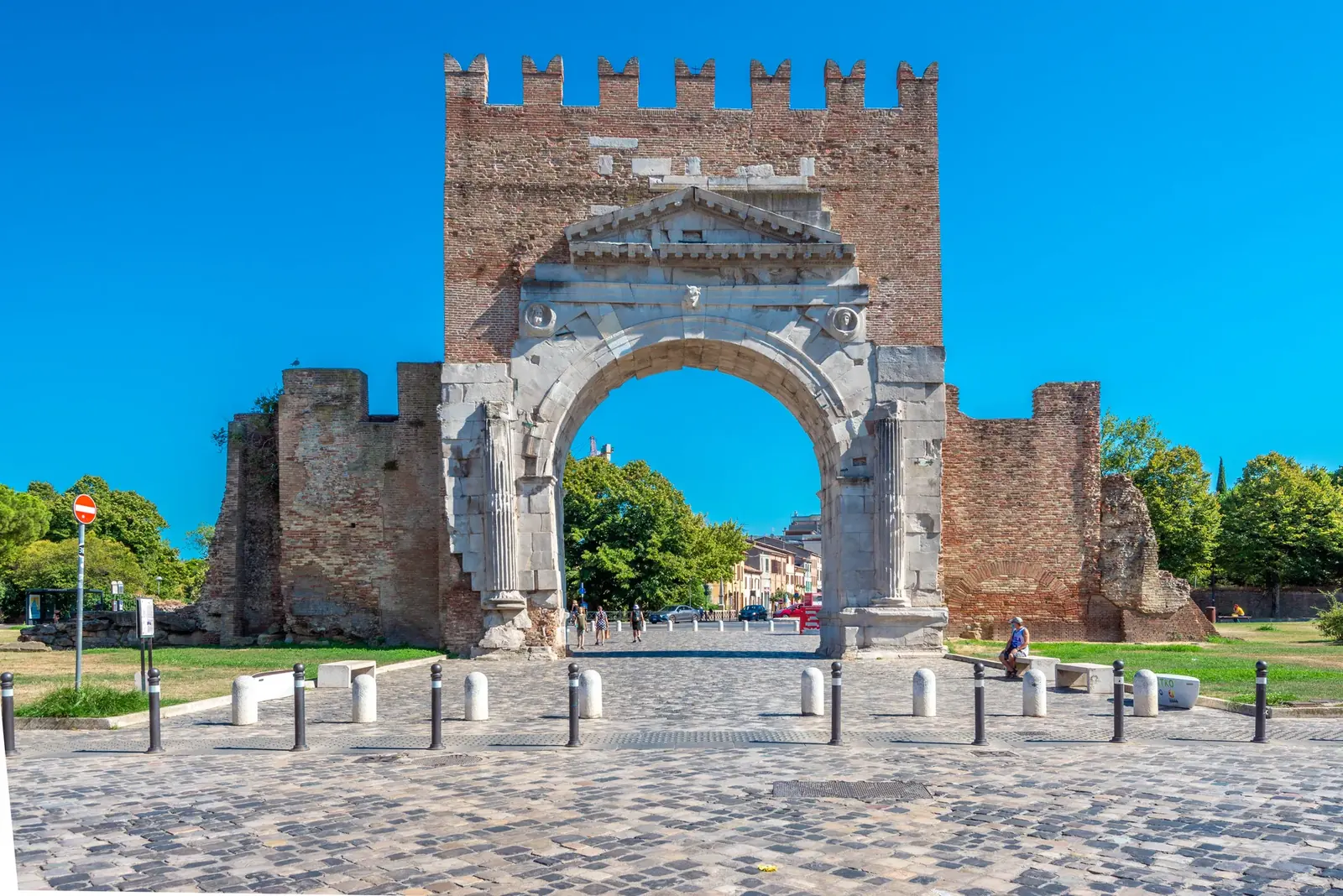 L'Arco di Augusto sotto il cielo azzurro, con la piazza lastricata davanti e i resti delle mura medievali ai lati