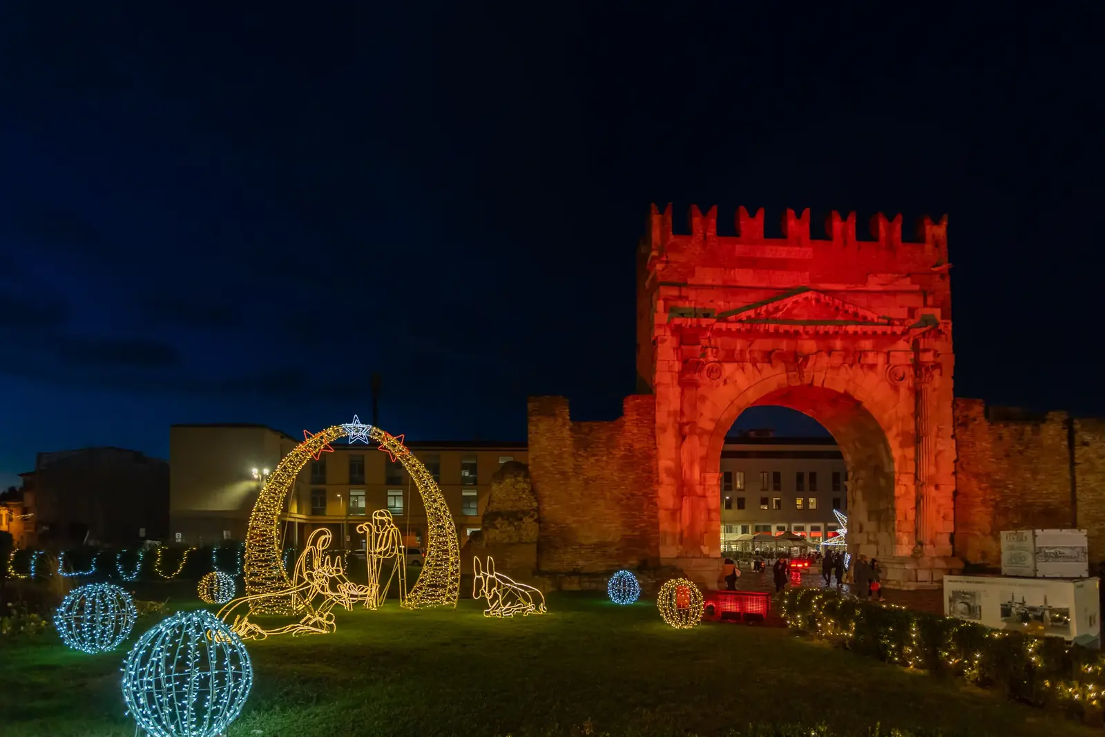 L'Arco di Augusto illuminato di rosso nelle sere di dicembre, con il presepe di luci e le sfere luminose sul prato