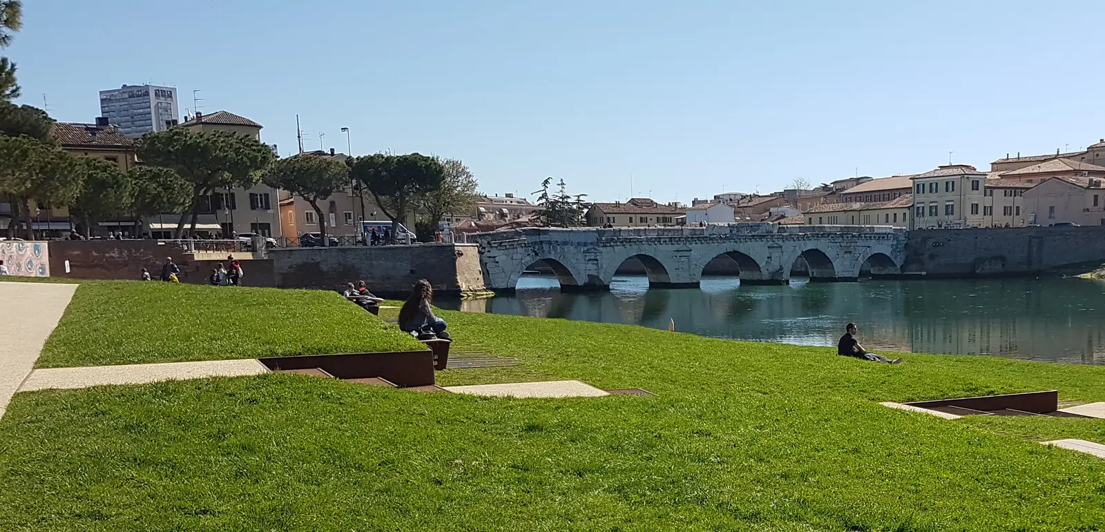Il prato della Piazza sull'Acqua a Rimini, con il Ponte di Tiberio e le case del Borgo sullo sfondo