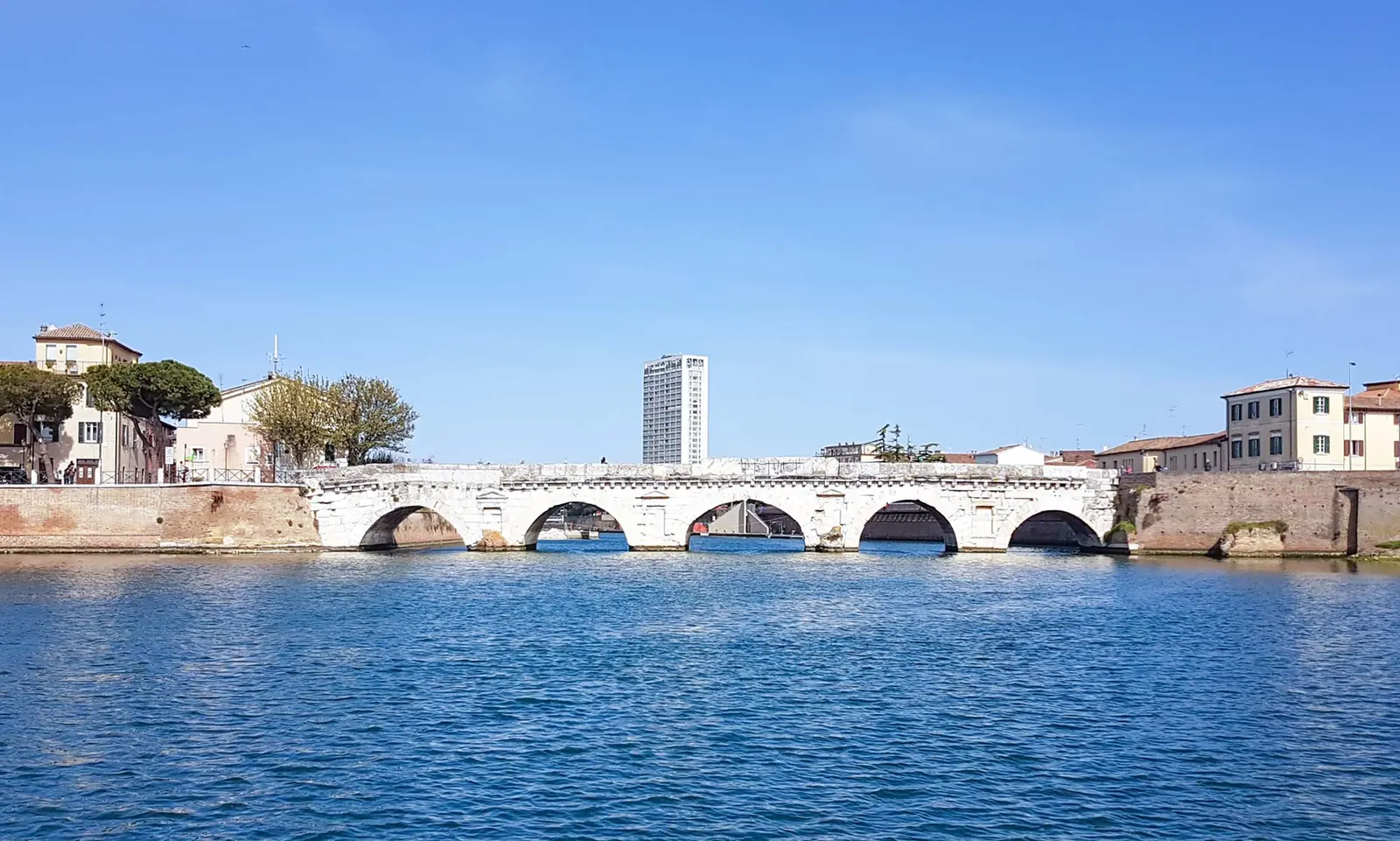 Il Ponte di Tiberio a Rimini visto dall'acqua, con le cinque arcate in pietra bianca e il grattacielo sullo sfondo