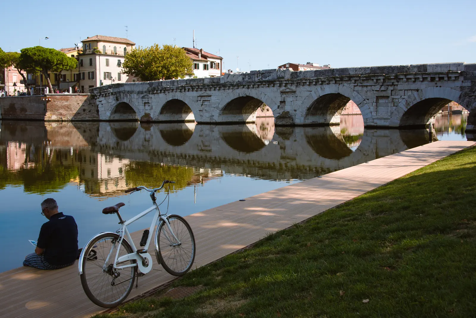 Il Ponte di Tiberio a Rimini con le arcate riflesse nel canale, una bicicletta bianca sulla passerella e il prato